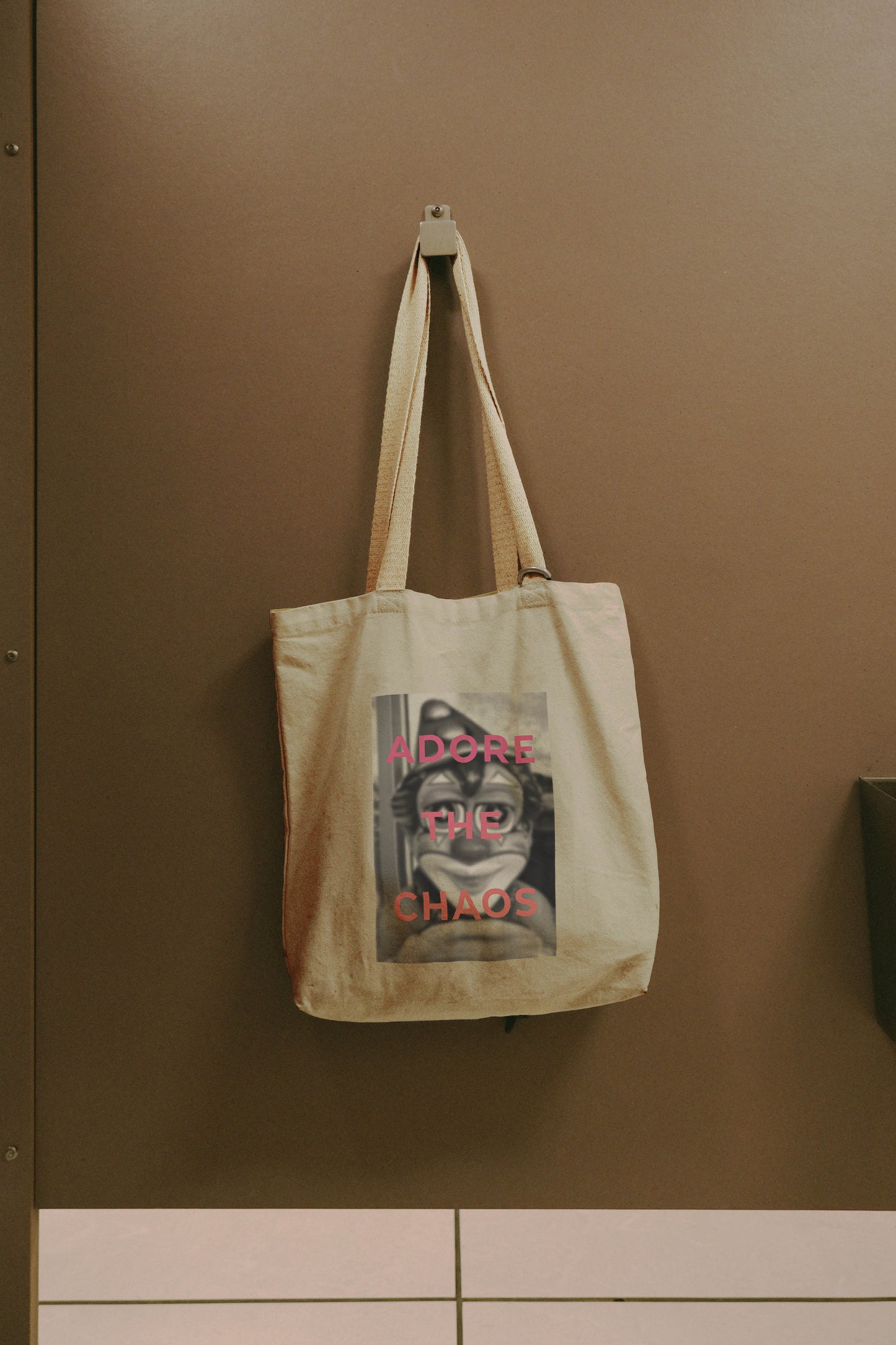 The Adore The Chaos - Tote Bag in beige natural cotton canvas hangs on a brown wall hook, featuring a monochrome image of a person with glasses and bold pink ADORE THE CHAOS text over the photo.
