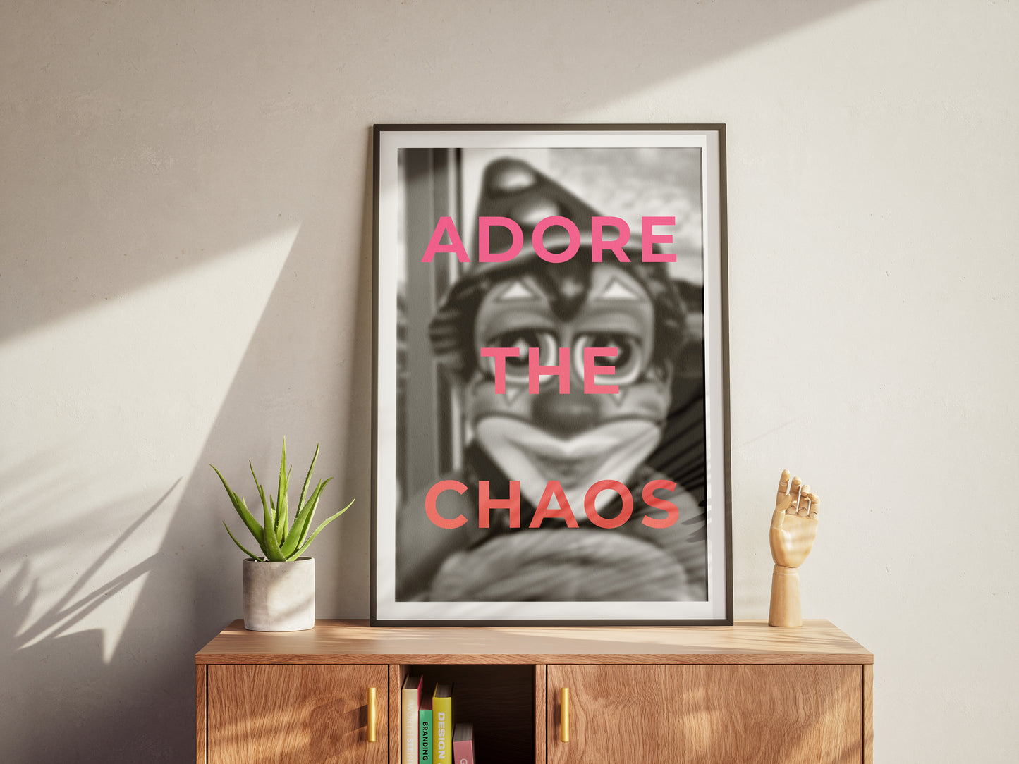The Adore The Chaos framed black-and-white art print with bold pink text sits on a wooden cabinet beside a potted plant and hand sculpture, adding modern flair to your decor as sunlight casts dramatic shadows on the wall.