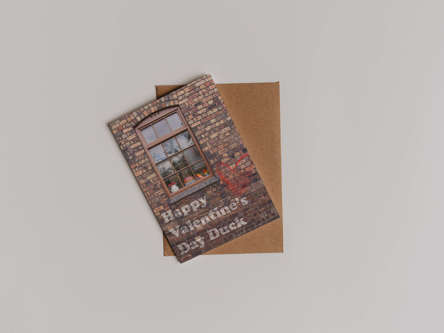 The Happy Valentines Day Duck Greetings Card features a brick wall and window design on eco-friendly cardstock, paired with a brown envelope. Perfect for sending love from Stoke-on-Trent.