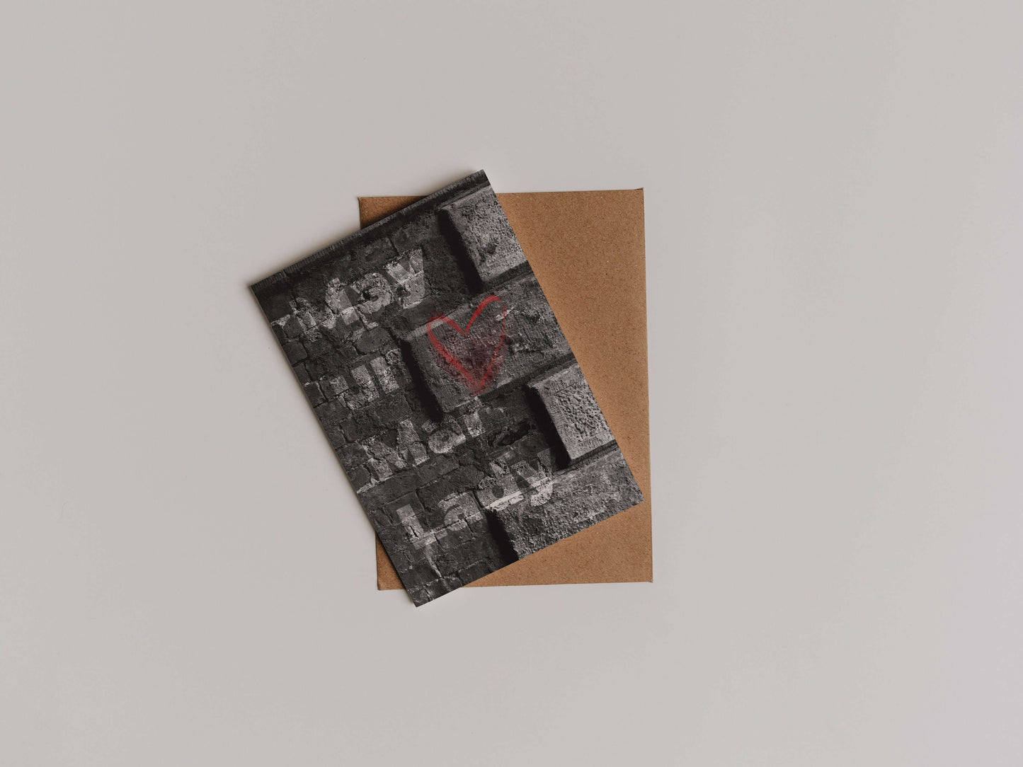 The May Un Mar Lady Greetings Card features a textured dark gray brick design with a red heart and Hey Hi You I See in white block letters, printed on eco-friendly cardstock. It comes with a brown envelope against a light background.