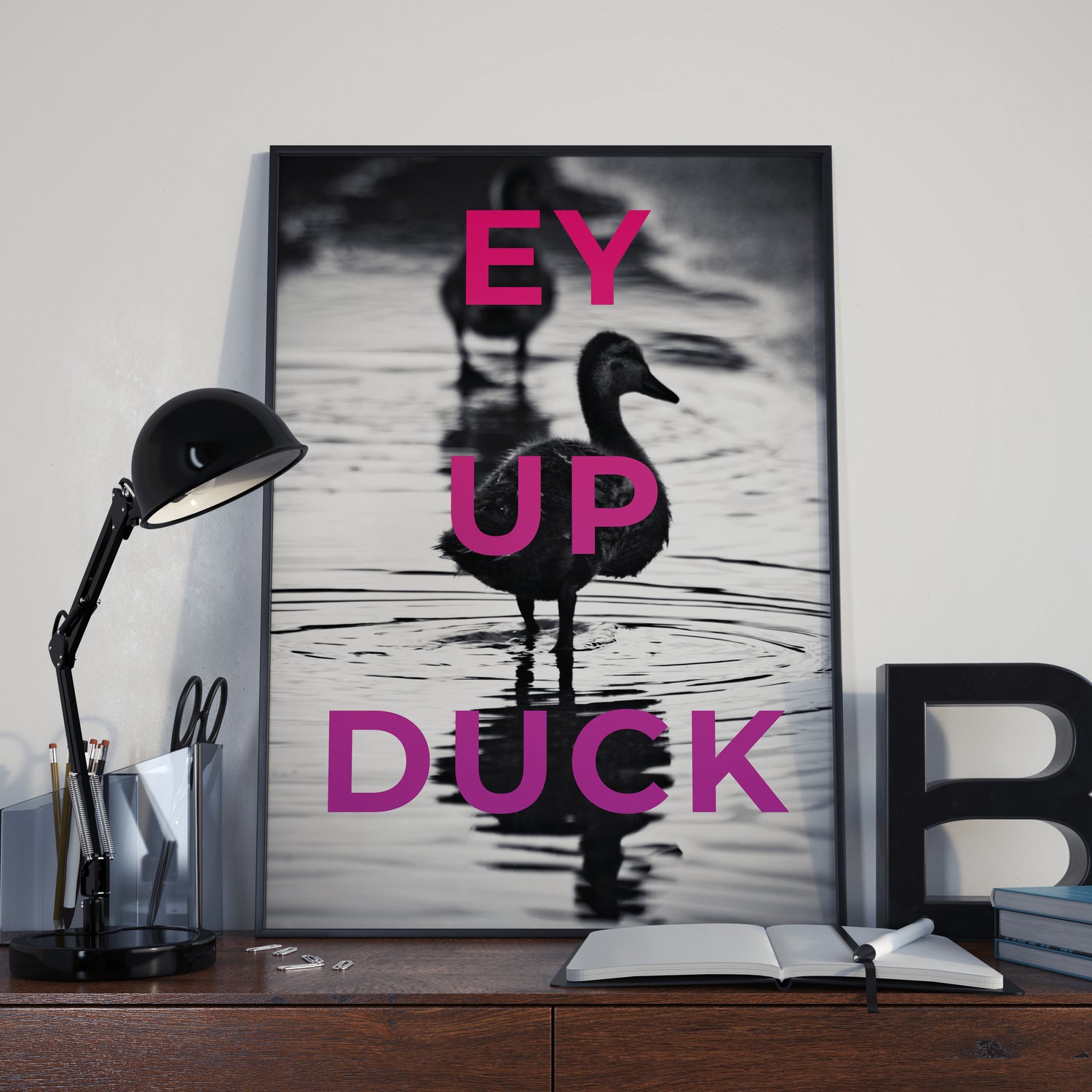 The Ey Up Duck framed art print features ducks in water with bold pink text over the image. Displayed on a desk, it adds playful regional charm alongside a lamp, stationery, books, and a decorative letter B.