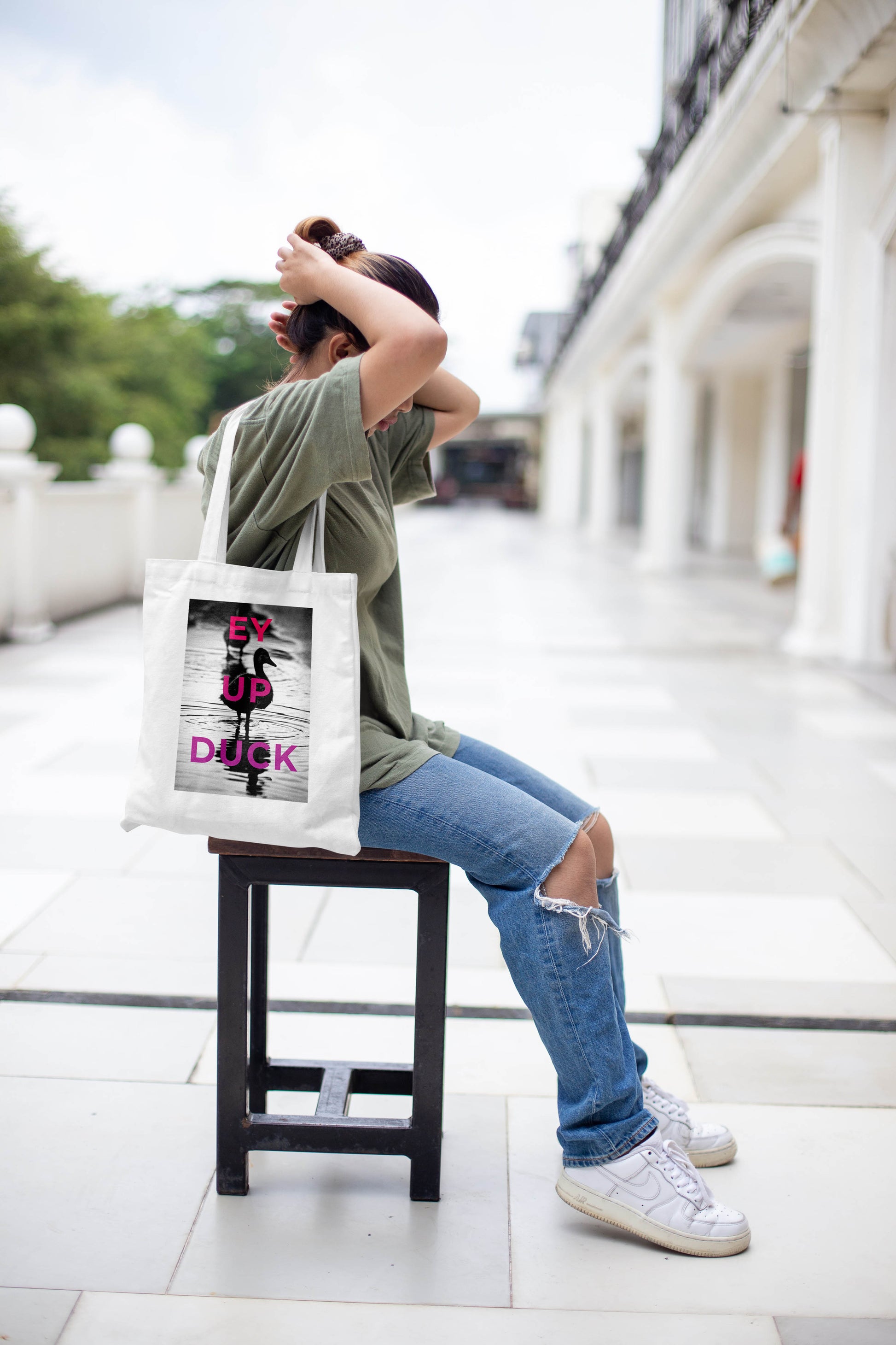 Outdoors, someone ties their hair while seated on a stool, dressed in a green shirt, ripped jeans, and white sneakers. They carry the Ey Up Duck - Tote Bag, its bold pink and black design adding a touch of Stoke charm to their look.