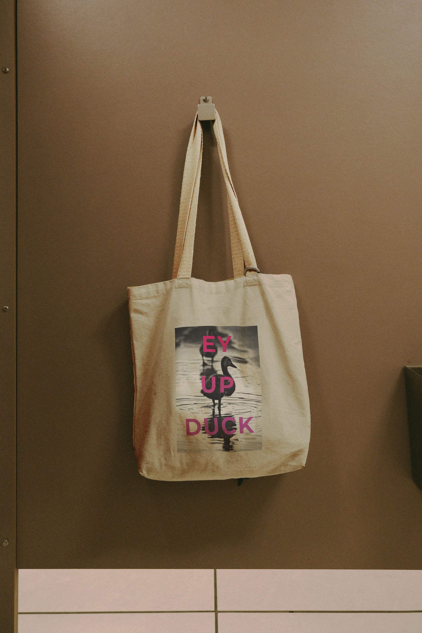 The Ey Up Duck - Tote Bag, featuring a duck on water and “Ey Up Duck” in bold pink letters, adds Stoke charm to your everyday carry; shown hanging on a brown wall hook.