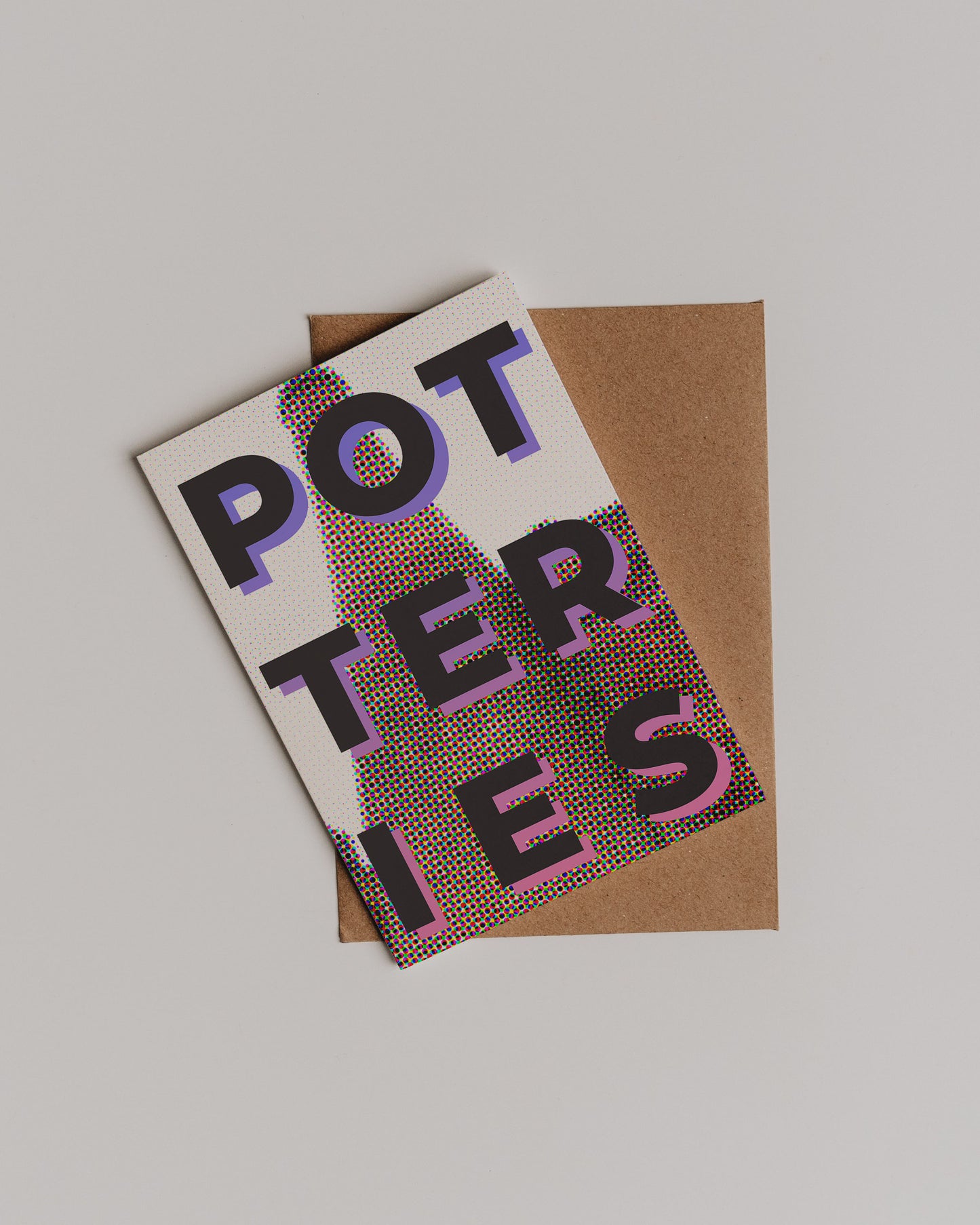 The Potteries Pop Art Greetings Card, featuring bold, colorful letters in traditional half tone print, lies diagonally on a brown envelope against a plain light background.