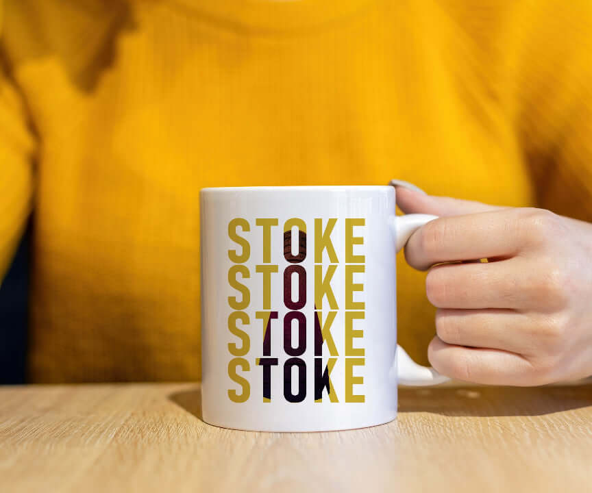 A pottery lover in a yellow sweater holds the Stoke Potbank Bottle Kiln Mug on a wooden table. The mug shows “STOKE” printed vertically five times in yellow and black, with one “TO” highlighted in red.