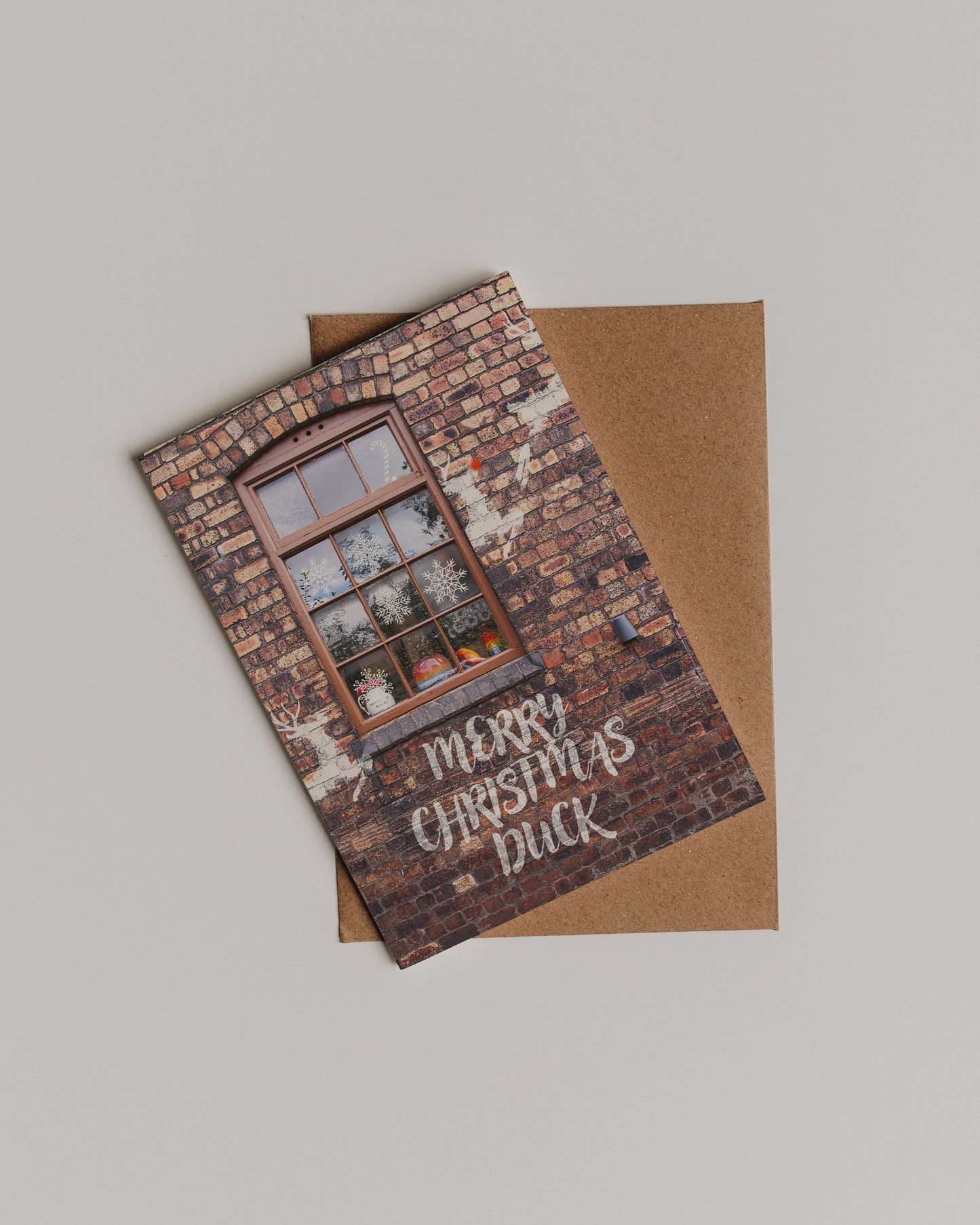 The Merry Christmas Duck Greetings Card shows a brick wall with a window, snowflakes, and festive décor. Merry Christmas Duck appears in white letters. The card sits atop a brown envelope, bringing Stoke-on-Trent cheer to your holiday.