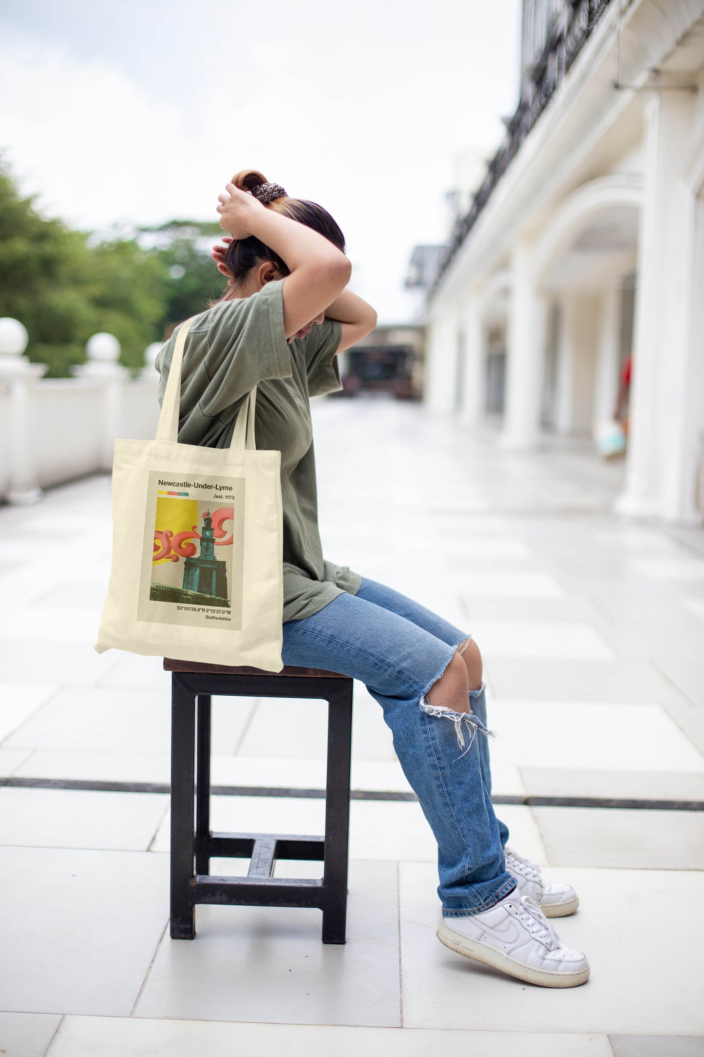 A person sits on a stool outdoors, tying their hair. They wear a green shirt, ripped jeans, white sneakers, and carry the Newcastle Under Lyme - Tote Bag with a colorful design. A white building with columns is in the background.