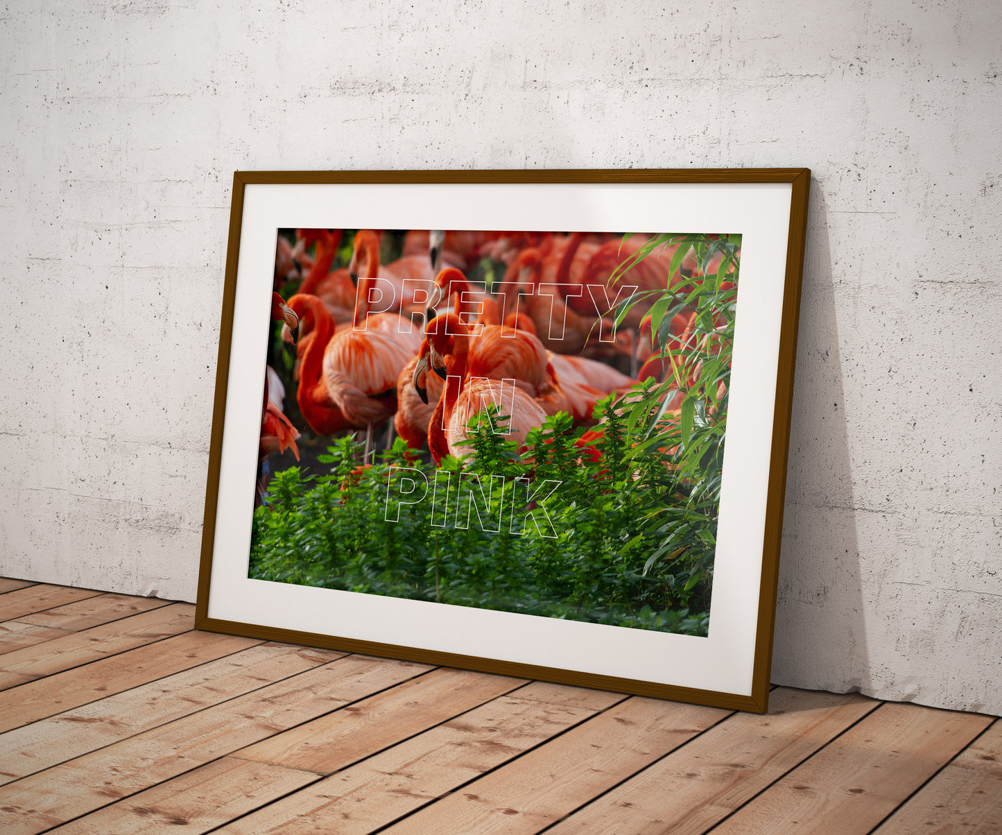 The Pretty In Pink print features a group of flamingos and bold text, framed and leaning against a white textured wall on a wooden floor. Green plants partially cover the premium card, adding playful energy to the décor.