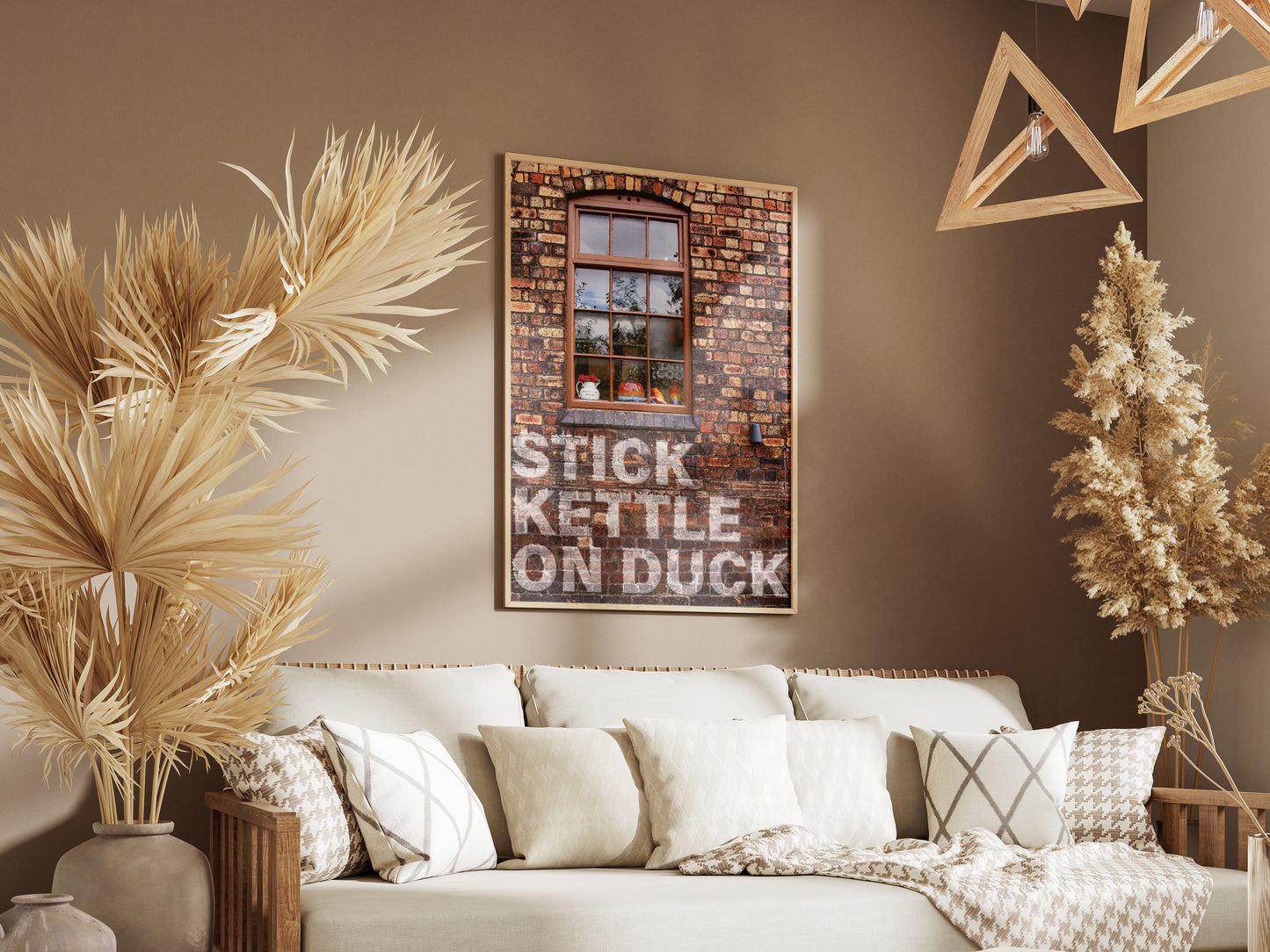 A cozy living room with neutral tones features a white sofa, patterned pillows, dried plants, geometric wooden lights, and framed Stick Kettle On wall art printed on high-quality card using HP Indigo technology below a window image.