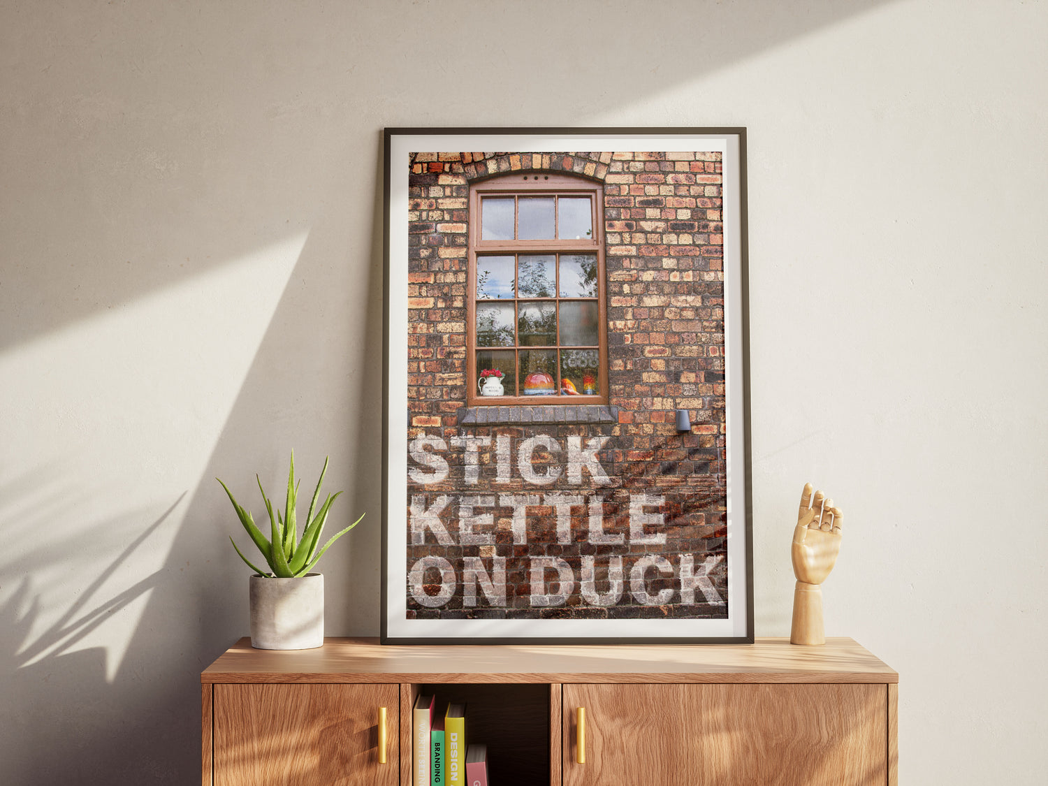 The Stick Kettle On framed poster, printed on premium card with HP Indigo technology, sits atop a wooden cabinet. The artwork features a brick wall and window scene, accompanied by an aloe plant and wooden hand sculpture nearby.