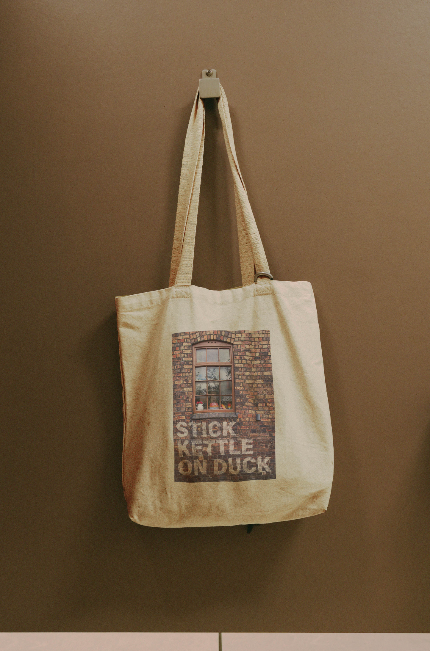 The Stick Kettle On - Tote Bag is a beige cotton canvas shopping bag printed with a brick wall, window, and the words STICK KETTLE ON DUCK, shown hanging on a hook against a brown wall.