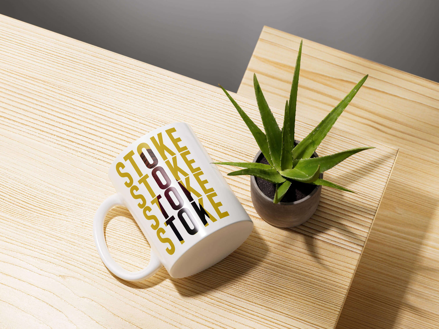 The Stoke Potbank Bottle Kiln Mug, featuring STOKE in yellow and black letters, lies on its side next to a small potted aloe—an ideal addition to any pottery enthusiast’s collection.