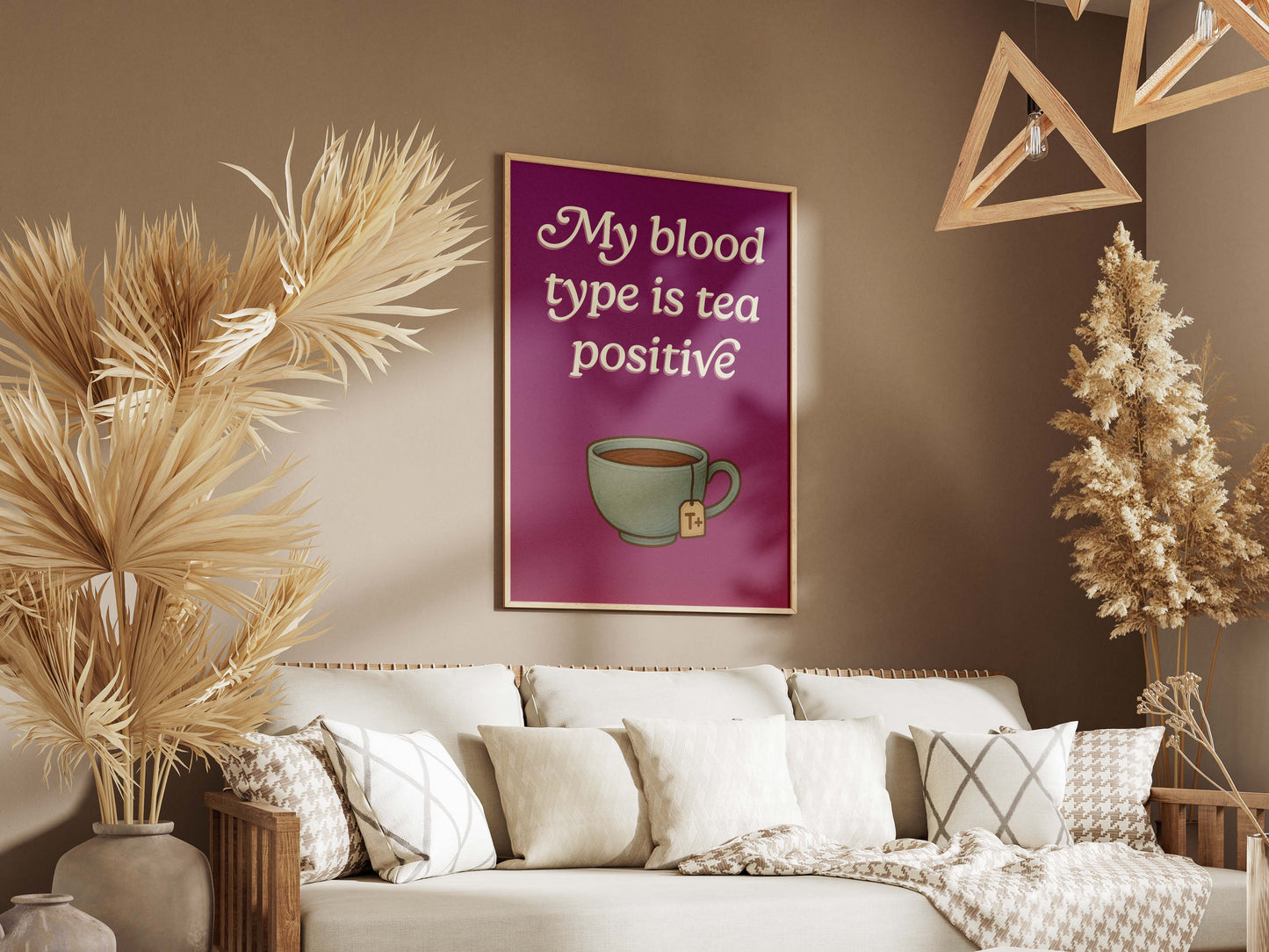A cozy living room with beige decor features the My Blood Type Is Tea Positive art print—showcasing a playful teacup—paired with a couch, patterned cushions, and dried plants. Perfect wall art for any tea lover.