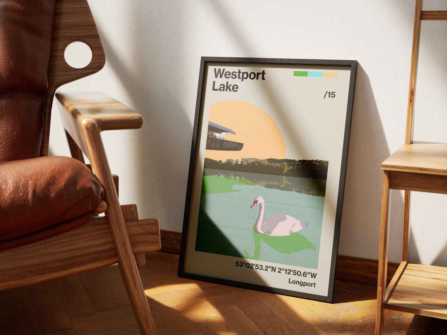 The Westport Lake Poster Print, shown framed and resting on a wall between two wooden chairs, features a swan on the lake, trees, a building, coordinates at the bottom, and sunlight in the background on high-quality paper.