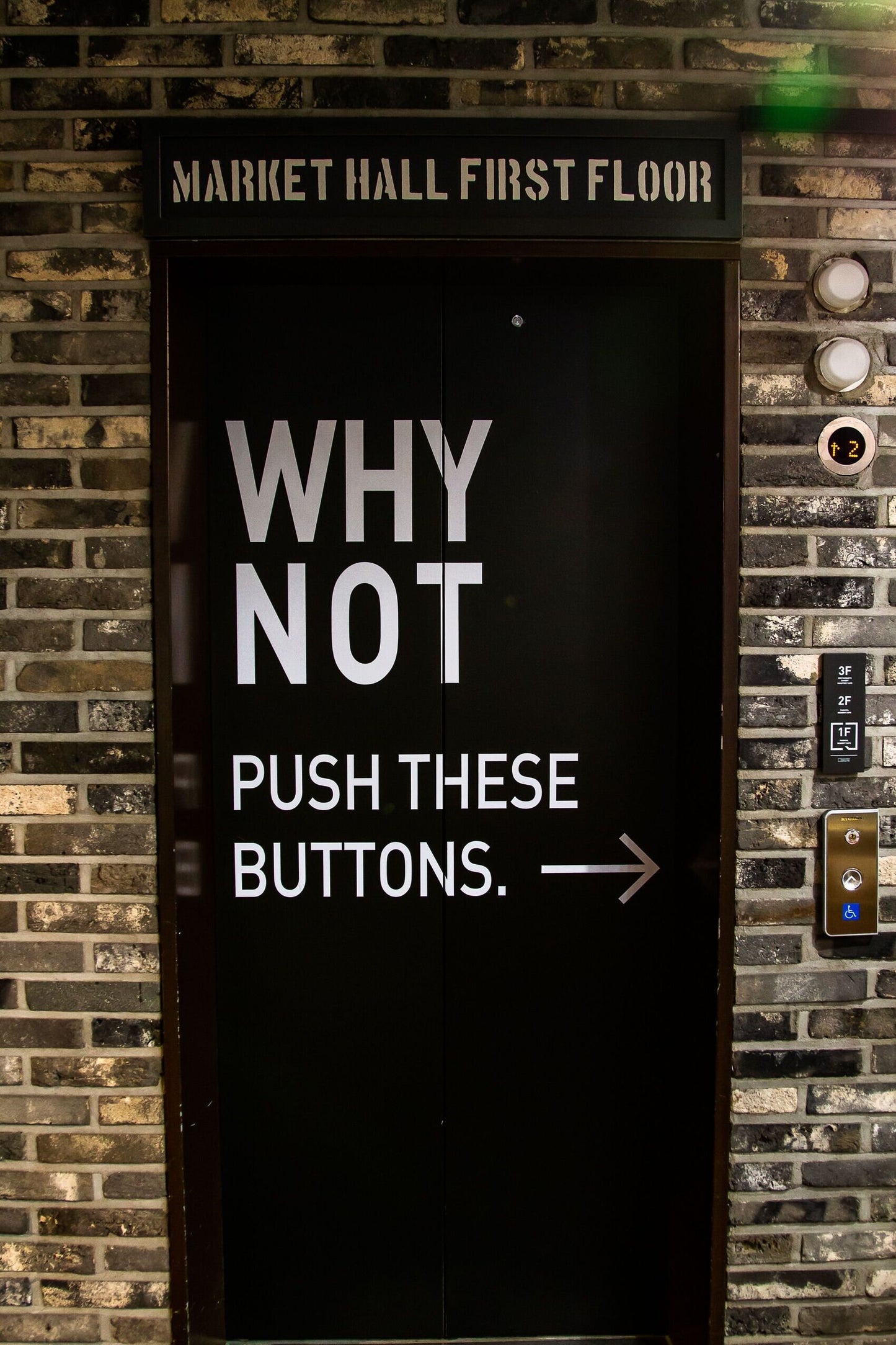 A sign reading MARKET HALL FIRST FLOOR hangs above an elevator door featuring the text Push These Buttons, with an arrow pointing right to a wall display that highlights the quality of paper used in Push These Buttons.
