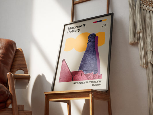 The Moorcroft Pottery Poster Print, framed and printed on high quality paper, leans against a wooden chair in sunlight, showcasing a bottle kiln illustration, geometric shapes, text, and Burslem coordinates in Stoke On Trent.